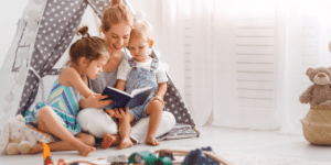 a mom sits with two girl toddlers on her lap while reading a book make reading at home fun Youthland academy