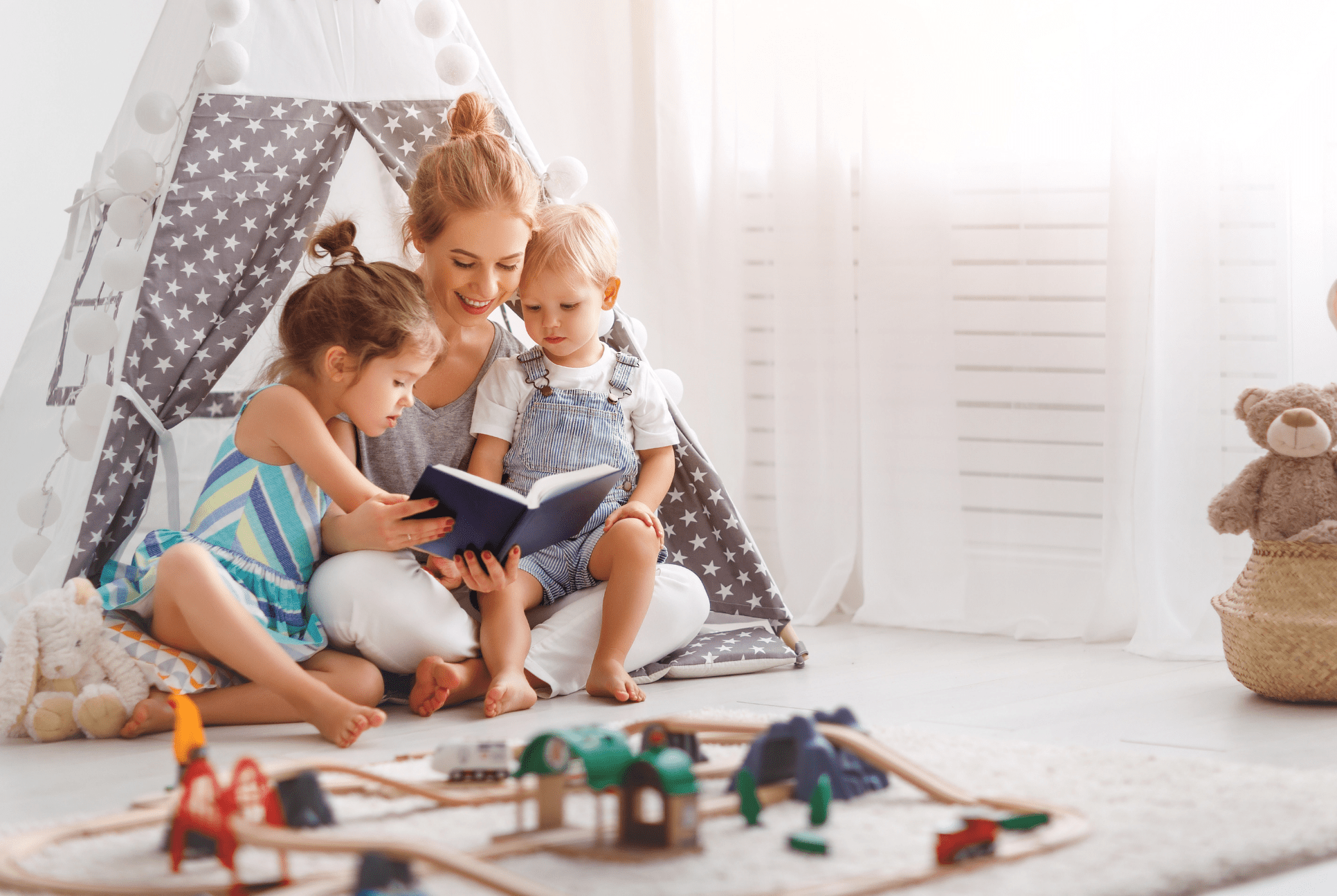 a mom sits with two girl toddlers on her lap while reading a book make reading at home fun Youthland academy