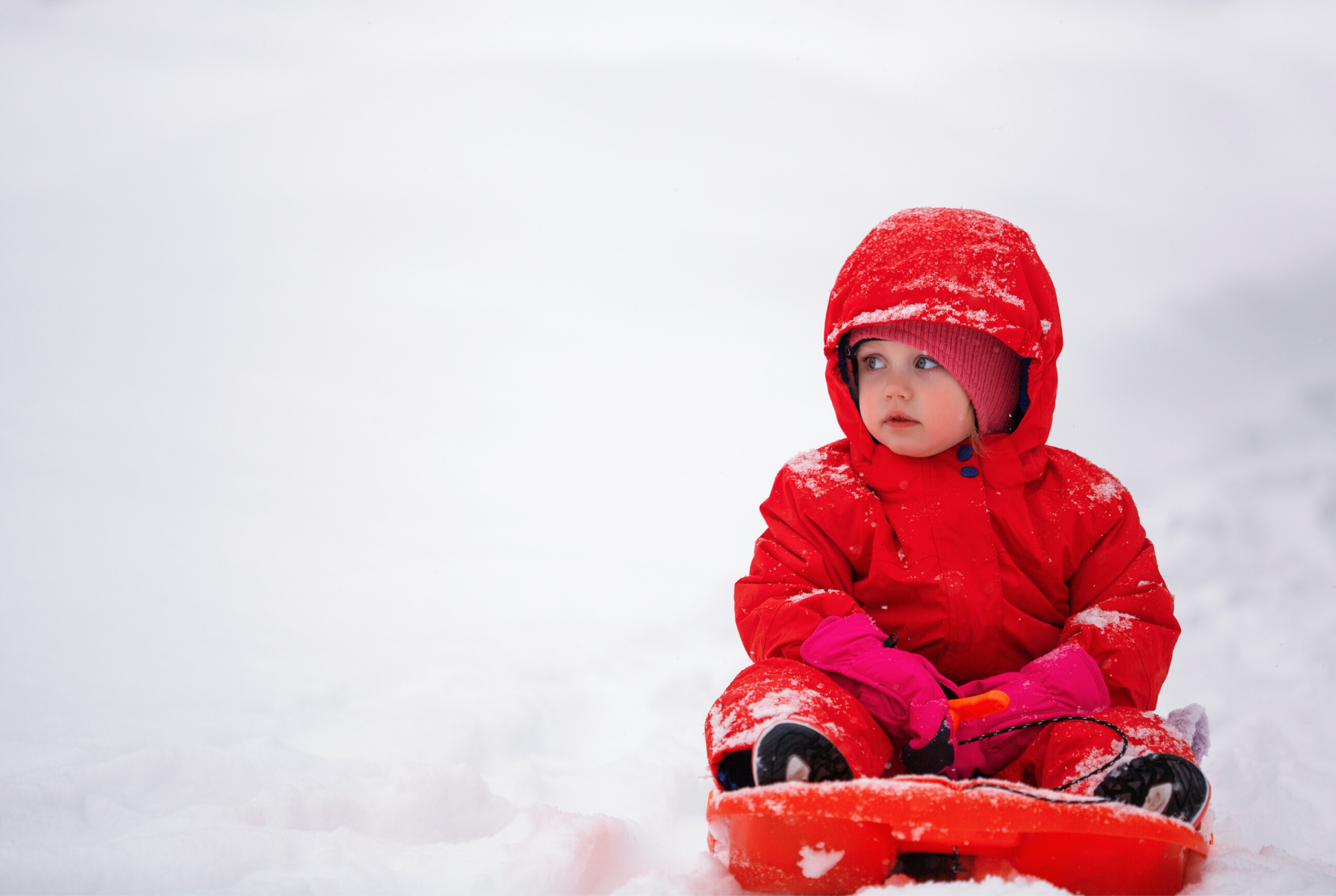 toddler in red snow suit sits on a red sled in the snow