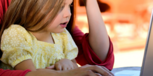 young girl sits on her parent's lap looking at a laptop